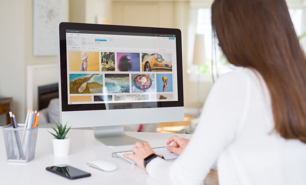 A woman working on a desktop computer displaying a digital asset management interface with various image files. The images are organized in a grid layout on the screen, representing efficient handling of visual content. The workspace includes a smartphone, a keyboard, and office supplies, symbolizing a modern, AI-powered digital asset management system.