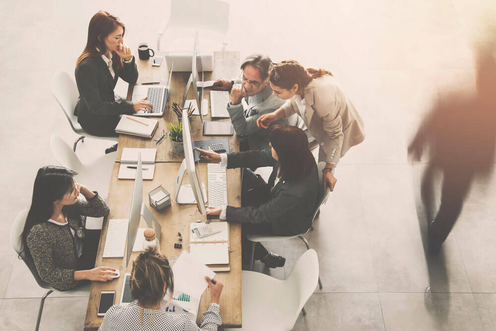 A group of people collaborating in an office setting, seated around a shared workspace with computers and documents. One person stands, pointing at a screen, while others engage in various tasks, symbolizing teamwork and digital asset management. The image conveys a busy, productive environment with a focus on digital tools.