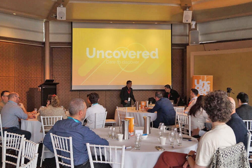 Deelnemers aan ronde tafels voeren discussies tijdens het WoodWing Uncovered APAC evenement op 6 november, met een helder presentatiescherm waarop het thema 'Dare to Discover' van het evenement wordt weergegeven.